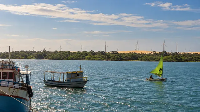 Barcos e um veleiro verde no Rio Jaguaribe, com eólicas ao fundo