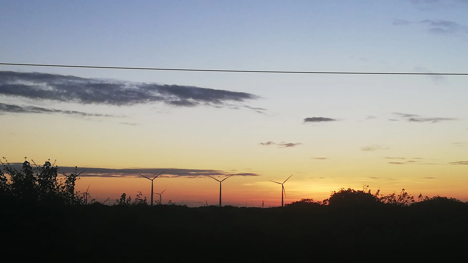 Silhueta das eólicas e da vegetação contra o céu alaranjado